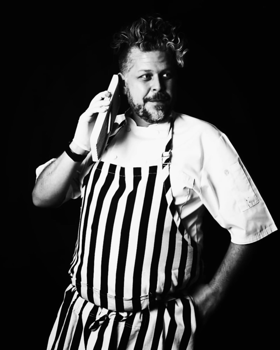 Black and white studio portrait in chef whites and striped apron, holding a mandoline like a telephone