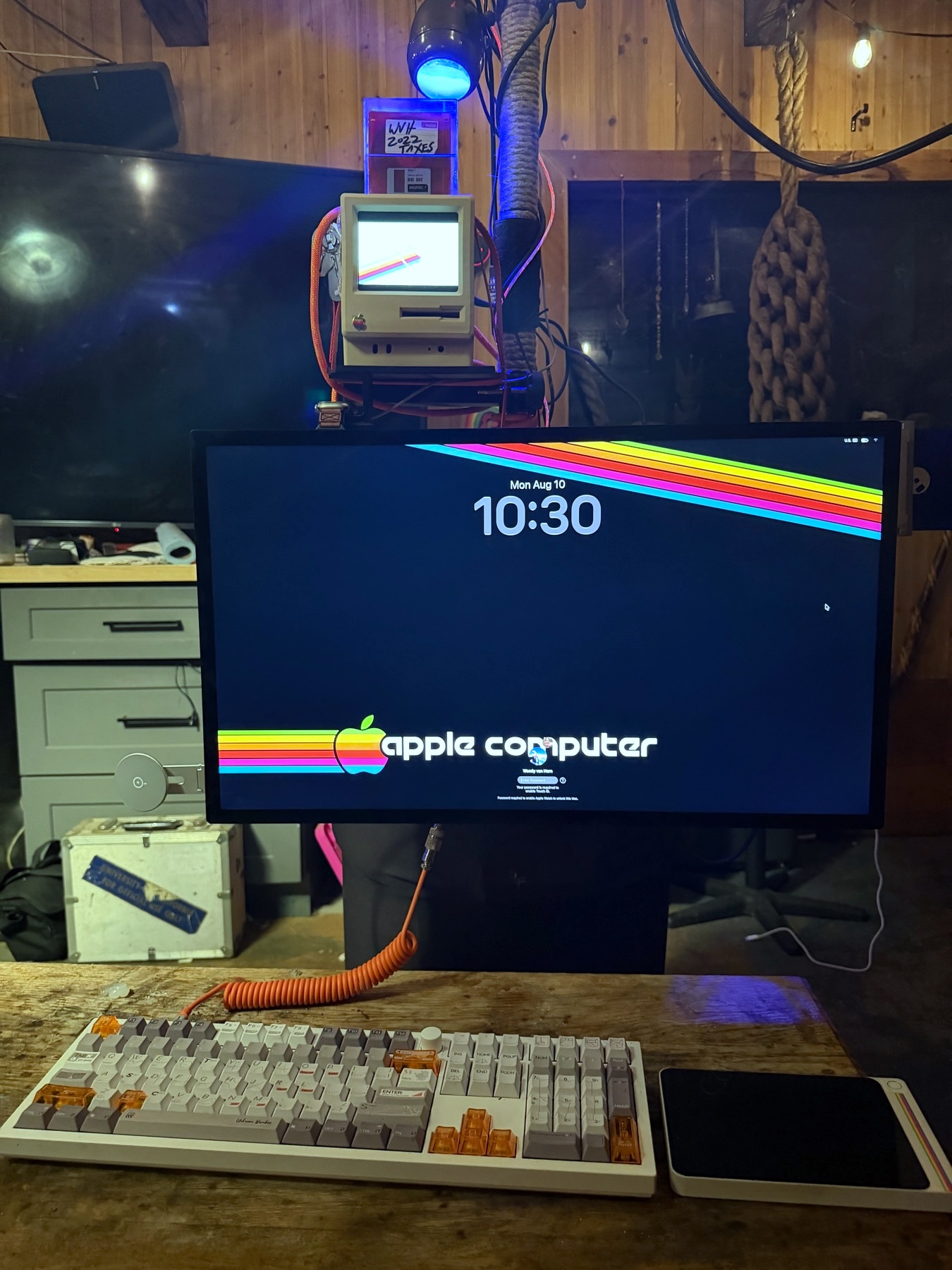 The Oracle at the desk - Apple Studio Display, a 3D-printed Macintosh Plus shell above it, and a Keychron keyboard on a reclaimed wood bench
